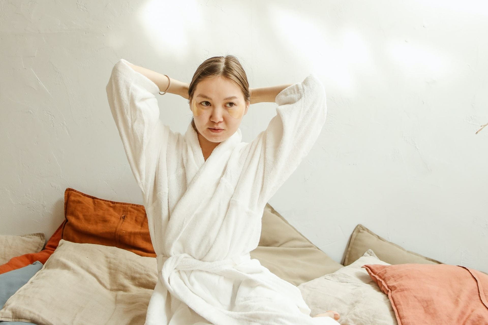 A woman sits on a bed in a robe while doing her hair.