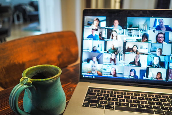 A coffee mug and a laptop on a table. On the laptop there is a Zoom meeting with blurred tiles of people's faces.